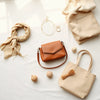 Neutrals accessories laid out on linen, including a leather bag, necklace, scarf, and earrings.