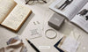 Flat lay of Styled Society xoxo minimalist jewelry including a silver bangle, heart studs, and a chain necklace on a desk with a notebook and fashion magazine.